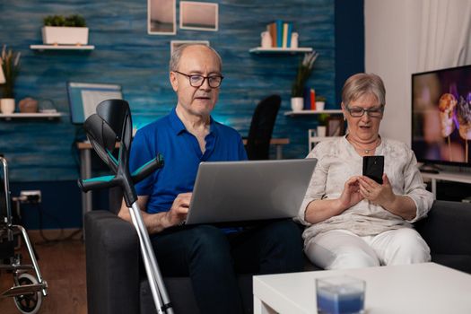 Senior family using modern technology devices laptop computer smartphone for browsing internet online while sitting on living room couch. Old people learning about digital gadget