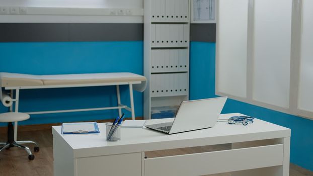 Nobody in medical cabinet space at healthcare facility. Empty doctors office designed with modern equipment, bed for consultation, laptop, documents, and stethoscope on white desk