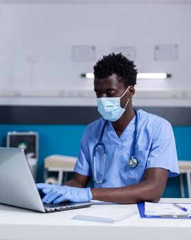 Black adult with nurse profession working at healthcare clinic using laptop and modern technology for medical system. African american man wearing uniform sitting at desk during pandemic