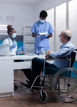 Professional african american team discussing healthcare diagnosis with disabled old patient. Black doctor and nurse talking to sick elder man with handicap sitting in wheelchair