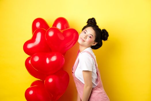 Valentines day concept. Stylish teenage asian girl posing near red hearts balloons, wear outfit for romantic date, standing happy on yellow background