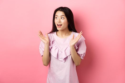 Excited asian girl clap hands and looking left, watching performance and applause, standing in dress on pink background