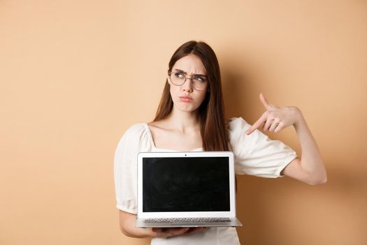 Suspicious frowning girl in glasses pointing at laptop screen, showing something strange online, standing on beige background