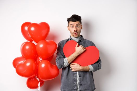 Surprised man holding Valentines heart card and saying wow, looking amazed at camera, receive secret confession on lovers day, standing near romantic balloons on white background