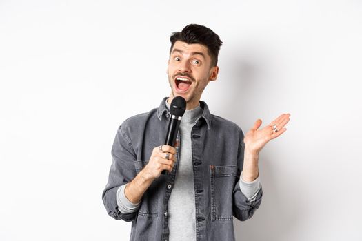 Excited stylish guy giving speech, talking in microphone, perform or sing karaoke, standing on white background