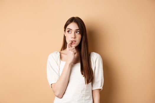 Nervous young girl biting fingernail and looking aside excited, making choice, thinking about something, standing on beige background