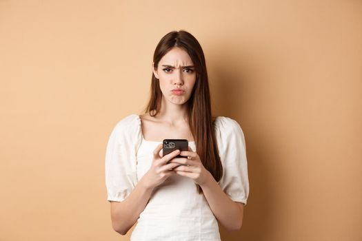 Disappointed girl with smartphone frowning, pucker lips upset, reading bad news on phone, standing on beige background
