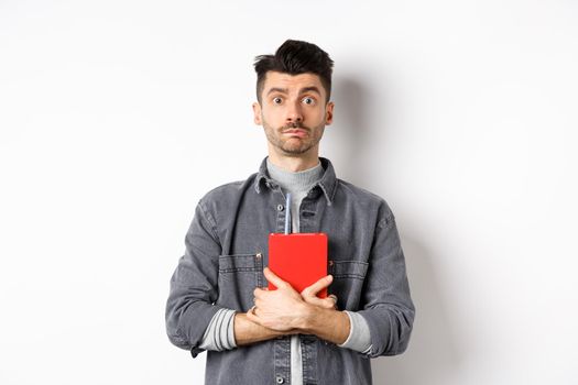 Cute and shy guy hugging his diary, holding red journal and look modest at camera, standing against white background