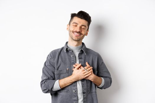 Romantic happy man holding hands on heart and smiling with closed eyes, dreaming of something, feeling nostalgic from sweet memories, standing on white background