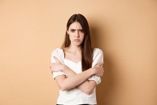 Offended woman hugging herself and frowning upset, feeling timid and defensive, standing on beige background