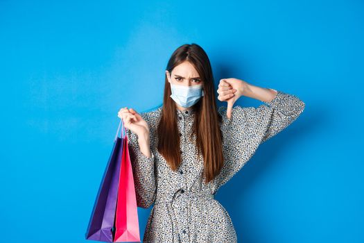 Covid-19, pandemic and lifestyle concept. Angry client in medical mask showing thumb down in dislike, frowning upset, holding shopping bags, blue background