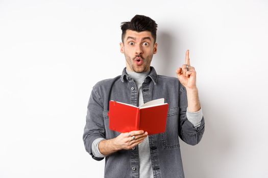 Excited man pitch an idea while reading planner or diary, holding red journal and gasping amazed, raising finger in eureka sign, standing on white background