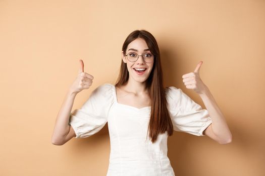 Excited young woman in glasses showing thumbs up and smiling, praising good optic store, approve and like eyewear, standing on beige background