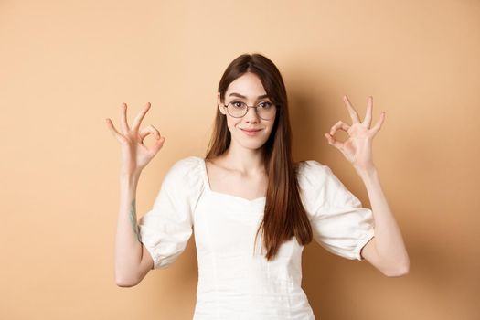 Satisfied female model in glasses show okay sign, looking pleased, agree and approve, standing on beige background