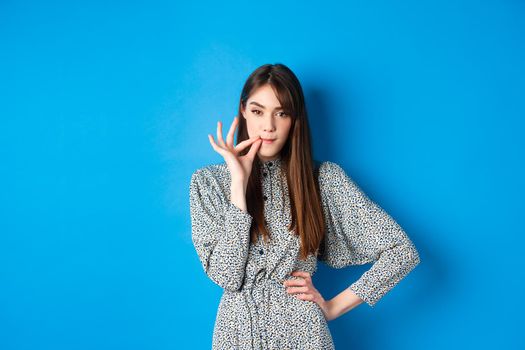 Serious woman promise to keep secret, seal lips, making zip gesture and looking at camera, standing in dress on blue background
