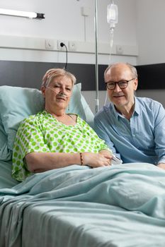 Retired hospitalized sick woman lying in bed with husband visitor sitting beside her