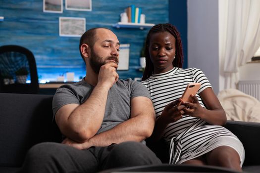 Interracial people using technology for entertainment at home. African american woman holding modern smartphone while caucasian man looking at digital gadget. Multi ethnic couple