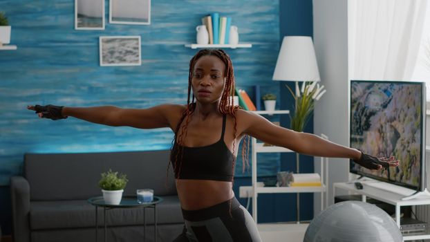 Slim athetic black woman wearing sportswear practicing leg stand posture during morning fitness workout in living room. Fit adult stretching body muscle enjoying healthy lifestyle