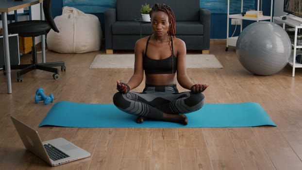 Flexible slim black woman sitting in lotus position on yoga map in living room