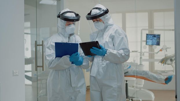 Dentist giving tablet to dental nurse before consultation appointment with patient. Stomatology doctors wearing ppe suits during pandemic while treating man for oral healthcare