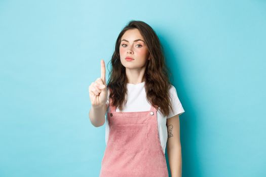 Rule number one. Serious young beautiful girl shaking finger in disapproval, scolding bad decision, make block or taboo gesture, standing against blue background