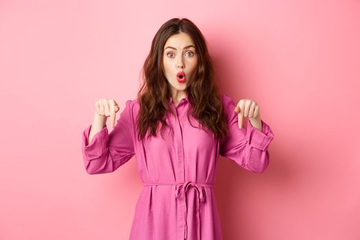 Stylish caucasian woman saying wow, pointing fingers down at impressive banner news, showing advertisement, staring amazed, standing against pink background