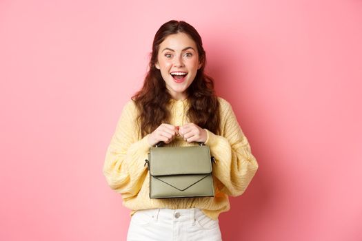 Image of happy young woman going on shopping, holding her purse and smiling excited, ready to go, standing against pink background