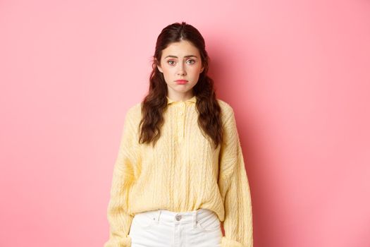 Sad cute woman looking innocent and silly at camera, standing against pink background