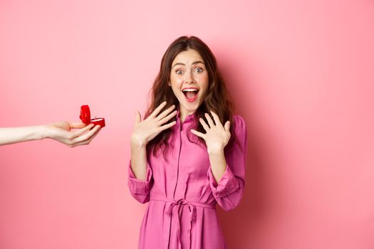 Portrait of surprised girlfriend scream of excitement as hand showing her engagement ring, recieve marriage proposal, look at camera amazed, standing over pink background