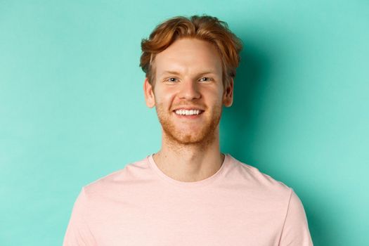 Close up of redhead bearded guy in pink t-shirt, smiling with white perfect teeth and looking at camera, standing over turquoise background.