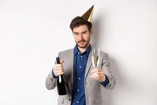 Celebration and holidays concept. Handsome man in suit and birthday hat giving glass of champagne, holding bottle alcohol, standing on white background.