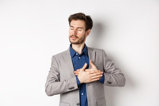 Romantic bearded man in stylish suit holding hands on heart, close eyes and remember something with tender feelings, standing nostalgic on white background