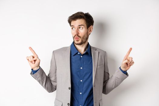 Indecisive male entrepreneur in suit pointing fingers sideways, choosing between two variants, looking at left logo pensive, standing on white background.