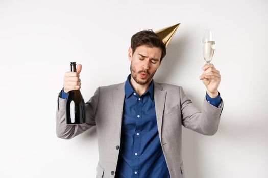 Celebration and holidays concept. Drunk guy dancing at party in birthday hat and suit, raising glass of champagne, having fun at event, white background.