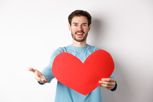 Handsome man in love making confession to you, pointing hand at camera, holding big red heart cutout on valentines day, singing romantic serenade, standing over white background