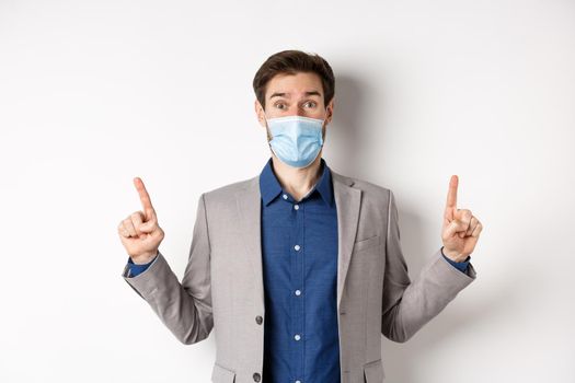 Covid-19, pandemic and business concept. Excited businessman in sterile medical mask and suit pointing fingers up, white background