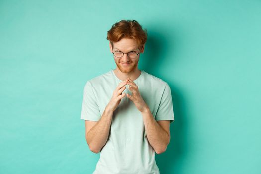Devious redhead man in glasses and t-shirt pitching an idea, steeple fingers and look from under forehead with sly and smug smile, standing over mint background