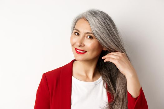 Beauty and haircare concept. Close up of elegant asian senior woman showing shiny and healthy grey hair, smiling and looking aside, white background