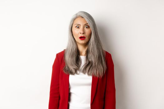 Business concept. Shocked senior female employer staring at camera speechless, standing in red blazer and makeup over white background