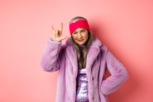 Funny and cool asian senior woman showing rock n roll gesture, looking sassy, standing over pink background