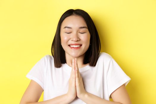 Close up of hopeful asian girl in white t-shirt, holding hands in pray, namste gesture and smiling, making wish or pleading, standing over yellow background.