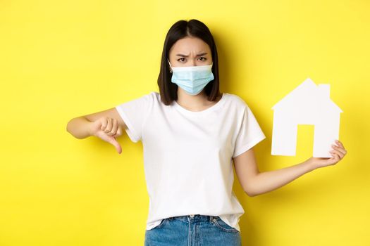 Covid-19 and real estate concept. Disappointed asian woman in medical mask, showing thumb down and paper house cutout, standing upset against yellow background