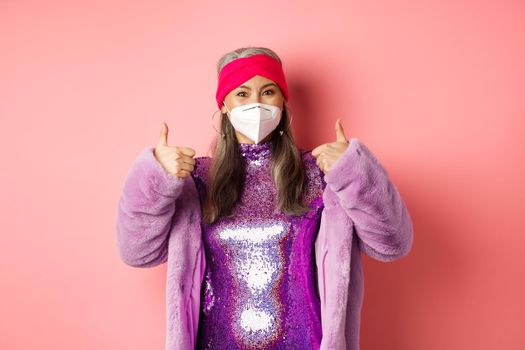 Covid-19, pandemic and fashion concept. Cheerful asian senior woman partying during coronavirus with respirator, showing thumbs-up, recommend wear face masks, pink background