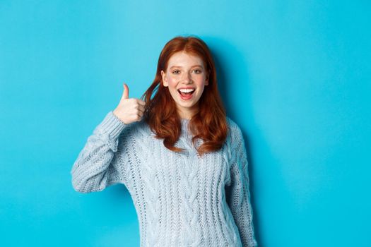 Cheerful redhead girl in sweater, showing thumb up in approval, like and praise product, standing over blue background