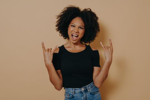 Young african woman with hands in rock n roll sign and shouting loudly, isolated on beige background