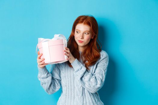Curious teen girl with red hair, shaking gift box and wonder what inside, standing over blue background