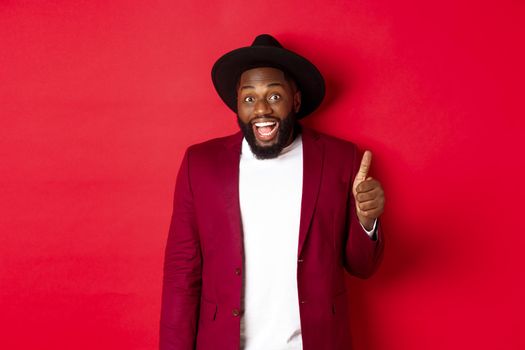 Christmas shopping and people concept. Handsome bearded Black man in party blazer showing thumb up, looking amazed and satisfied, red background.
