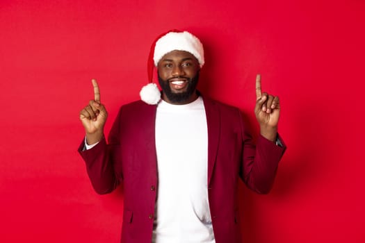 Christmas, party and holidays concept. Handsome african american man in santa hat smiling, pointing fingers up and showing advertisement, standing over red background.