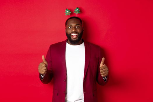 Merry Christmas. Happy african american man celebarting New Year, wearing funny party headband and showing thumb up, like and praise something, standing over red backgrond