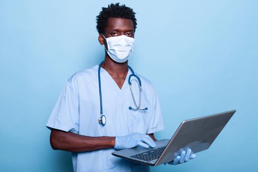 Healthcare assistant with laptop in hand wearing face mask. Medical nurse with uniform and gloves for coronavirus pandemic protection holding device. Specialist with stethoscope